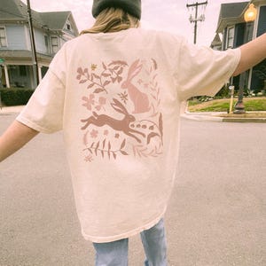 Pu&ograve; includere: Una t-shirt color crema con un design floreale rosa con due conigli e foglie. Il design &egrave; sul retro della t-shirt.