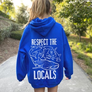 以下が含まれることがあります： 白のテキストとグラフィックが入ったロイヤルブルーのパーカー。「RESPECT THE LOCALS」の文字と、サメとクラゲの白いイラストがパーカーの背面に描かれています。