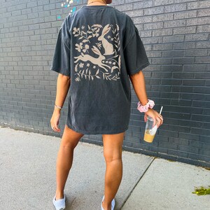 Pu&ograve; includere: Una maglietta oversize di colore grigio scuro con una stampa grafica bianca di tre conigli e disegni floreali sul retro.