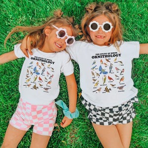 May include: Two children wearing white t-shirts with the text "The Study of Birds Ornithology" and a variety of bird illustrations. The girl on the left is wearing pink and white checked shorts and the girl on the right is wearing black and white checked shorts. Both girls are wearing white sunglasses.