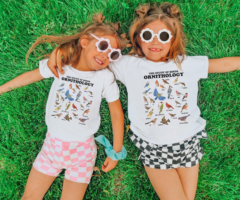 May include: Two children wearing white t-shirts with the text "The Study of Birds Ornithology" and a variety of bird illustrations. The girl on the left is wearing pink and white checked shorts and the girl on the right is wearing black and white checked shorts. Both girls are wearing white sunglasses.