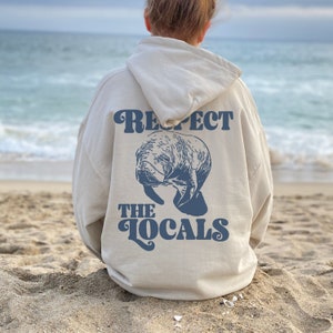 May include: A cream-colored hoodie with a blue manatee graphic and the text "Respect the Locals" on the back. The hoodie is worn by a person sitting on a sandy beach.
