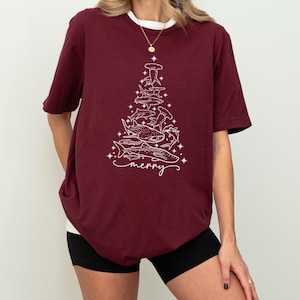 K&ouml;nnte beinhalten: Burgunderfarbenes T-Shirt mit einer wei&szlig;en Grafik eines Weihnachtsbaums aus Haien und dem Wort "merry". Das Shirt hat einen wei&szlig;en Kragen und kurze &Auml;rmel. Das Design zeigt verschiedene Haiarten.