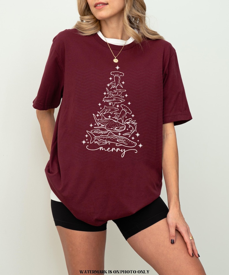 K&ouml;nnte beinhalten: Burgunderfarbenes T-Shirt mit einer wei&szlig;en Grafik eines Weihnachtsbaums aus Haien und dem Wort "merry". Das Shirt hat einen wei&szlig;en Kragen und kurze &Auml;rmel. Das Design zeigt verschiedene Haiarten.
