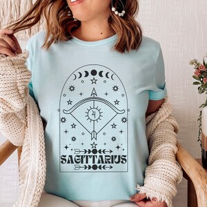 May include: A light blue t-shirt with a black graphic design of a Sagittarius zodiac symbol. The design features an arch with stars, moons, and an arrow. The text "Sagittarius" is below the arch.