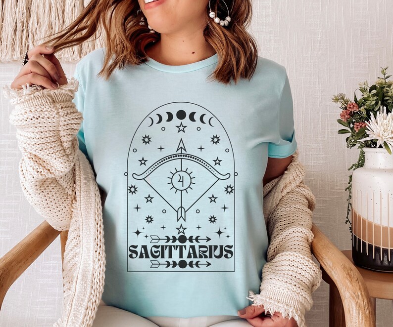 May include: A light blue t-shirt with a black graphic design of a Sagittarius zodiac symbol. The design features an arch with stars, moons, and an arrow. The text "Sagittarius" is below the arch.