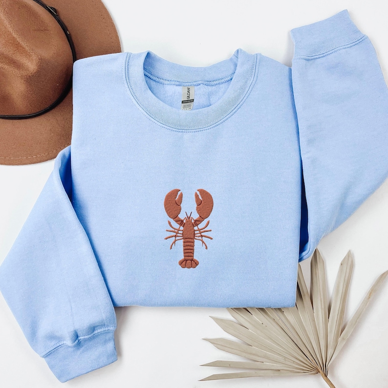 Pode incluir: Uma camisa de moletom azul claro com um lagosta bordada marrom na frente.