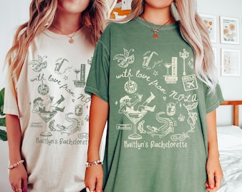 May include: Two t-shirts, one cream and one green, with a graphic design. The design includes the text "with love from NOLA" and "Kaitlyn's Bachelorette", along with illustrations of a cocktail, a saxophone, and a disco ball.