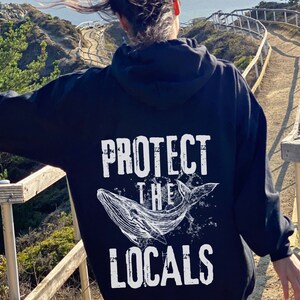 Może przedstawiać: Czarna bluza z kapturem z białym napisem "PROTECT THE LOCALS" i ilustracją wieloryba. Projekt ma postarzaną, vintage estetykę. Bluza jest noszona na zewnątrz.