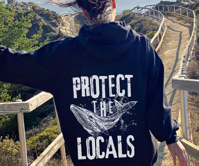 Może przedstawiać: Czarna bluza z kapturem z białym napisem "PROTECT THE LOCALS" i ilustracją wieloryba. Projekt ma postarzaną, vintage estetykę. Bluza jest noszona na zewnątrz.