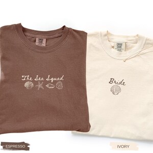 May include: An espresso brown t-shirt with the embroidered text The Sea Squad and four seashell icons, alongside an ivory t-shirt with the embroidered text Bride and a single seashell icon. Both shirts feature a Comfort Colors brand neck label.