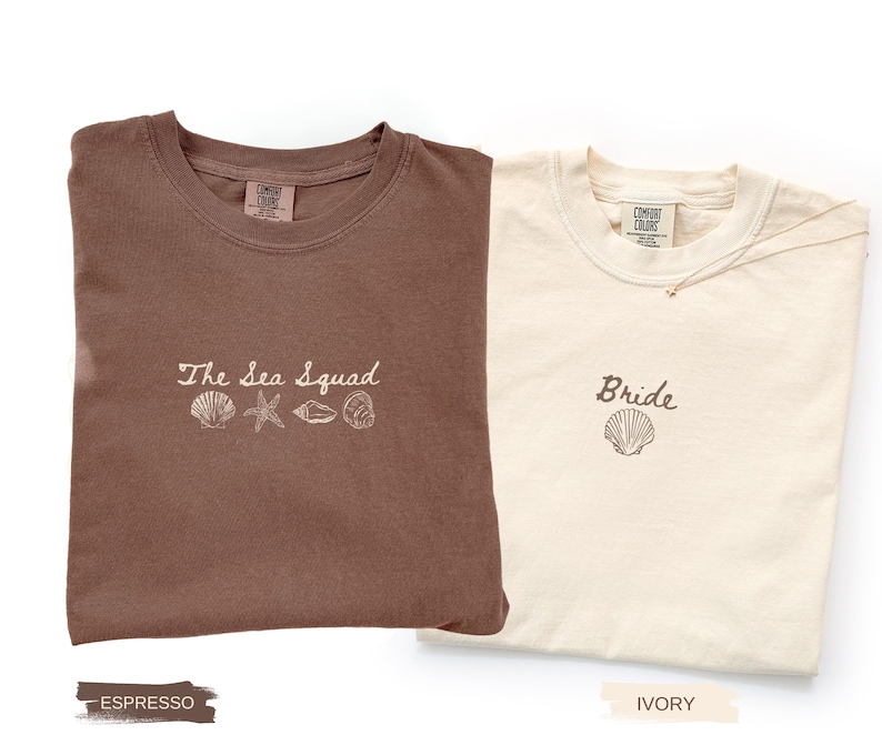 May include: An espresso brown t-shirt with the embroidered text The Sea Squad and four seashell icons, alongside an ivory t-shirt with the embroidered text Bride and a single seashell icon. Both shirts feature a Comfort Colors brand neck label.