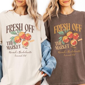 May include: Two t-shirts, one cream and one brown, featuring the text "FRESH OFF THE MARKET" above a peach graphic. The text "Hanah's Bachelorette Savannah GA" is below. The cream shirt has a light blue denim jacket.