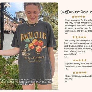 May include: Dark gray t-shirt with the text "BACH CLUB" in yellow and orange, featuring a fruit graphic. The shirt also includes the text "FRESH OFF THE MARKET" and "Katej's Bachelorette Savannah Georgia".