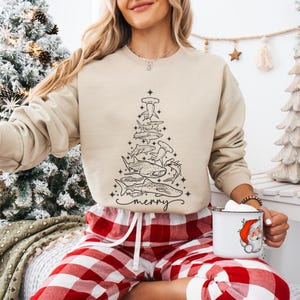K&ouml;nnte beinhalten: Beiges Sweatshirt mit einer schwarzen Strichzeichnung eines Weihnachtsbaums aus verschiedenen Haiarten. Das Wort "merry" steht in Schreibschrift am unteren Rand des Baumes. Die Person tr&auml;gt eine rot-wei&szlig; karierte Pyjamahose.