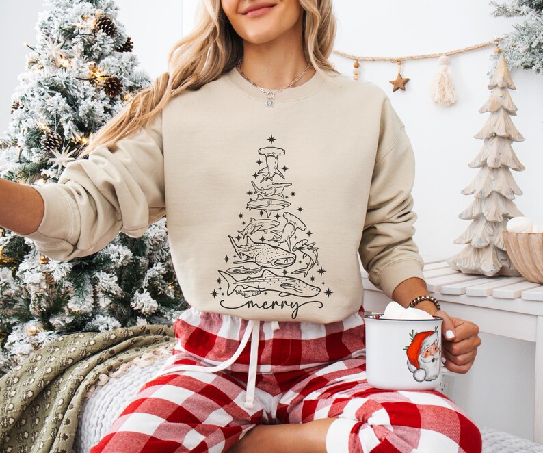 K&ouml;nnte beinhalten: Beiges Sweatshirt mit einer schwarzen Strichzeichnung eines Weihnachtsbaums aus verschiedenen Haiarten. Das Wort "merry" steht in Schreibschrift am unteren Rand des Baumes. Die Person tr&auml;gt eine rot-wei&szlig; karierte Pyjamahose.