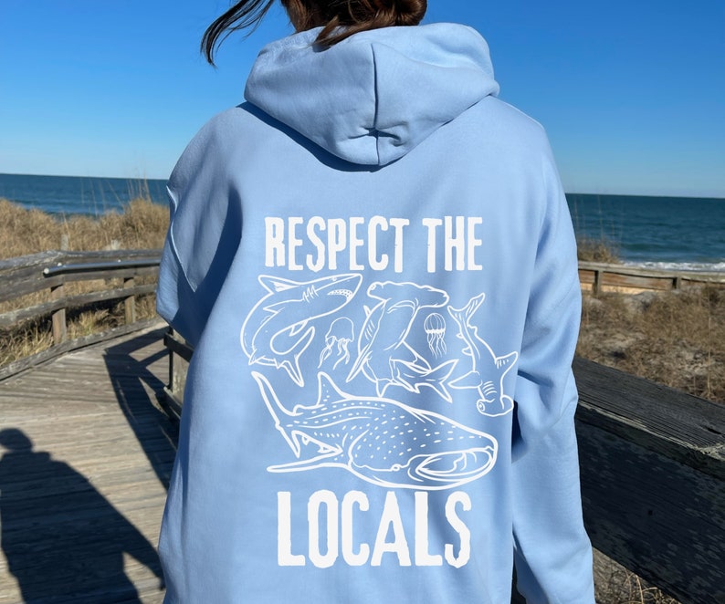 以下が含まれることがあります： 様々なサメの白いグラフィックと「RESPECT THE LOCALS」の文字が入った、水色のフード付きスウェットシャツ。フードと長袖が付いています。デザインは背面にプリントされています。