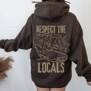 以下が含まれることがあります： 長袖とフード付きの濃い茶色のパーカー。背中には「RESPECT THE LOCALS」の文字と、サメやクラゲのベージュ色のグラフィックが描かれています。カジュアルなフィット感のパーカーです。