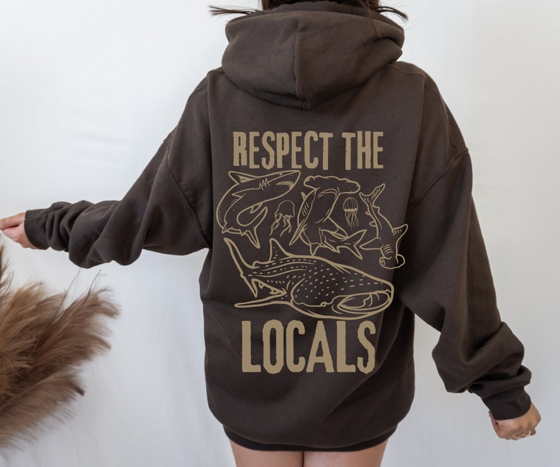 以下が含まれることがあります： 長袖とフード付きの濃い茶色のパーカー。背中には「RESPECT THE LOCALS」の文字と、サメやクラゲのベージュ色のグラフィックが描かれています。カジュアルなフィット感のパーカーです。