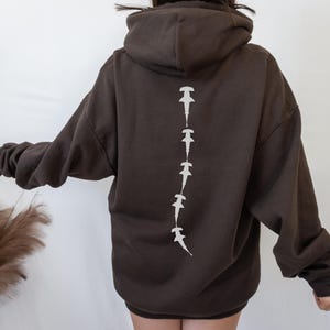 May include: A brown hoodie with a white hammerhead shark design on the back. The sharks are arranged in a vertical line down the center of the back.