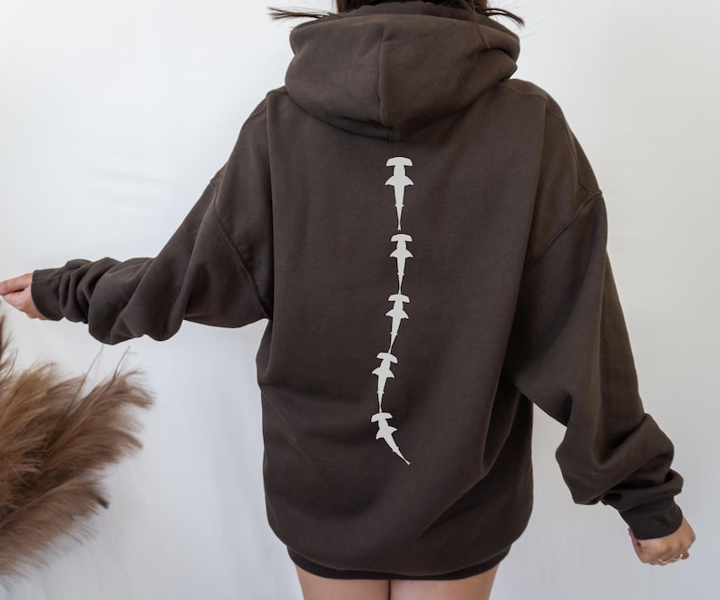 May include: A brown hoodie with a white hammerhead shark design on the back. The sharks are arranged in a vertical line down the center of the back.