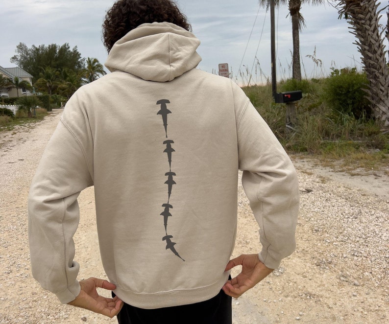 May include: A beige hoodie with a line of hammerhead shark silhouettes printed on the back.