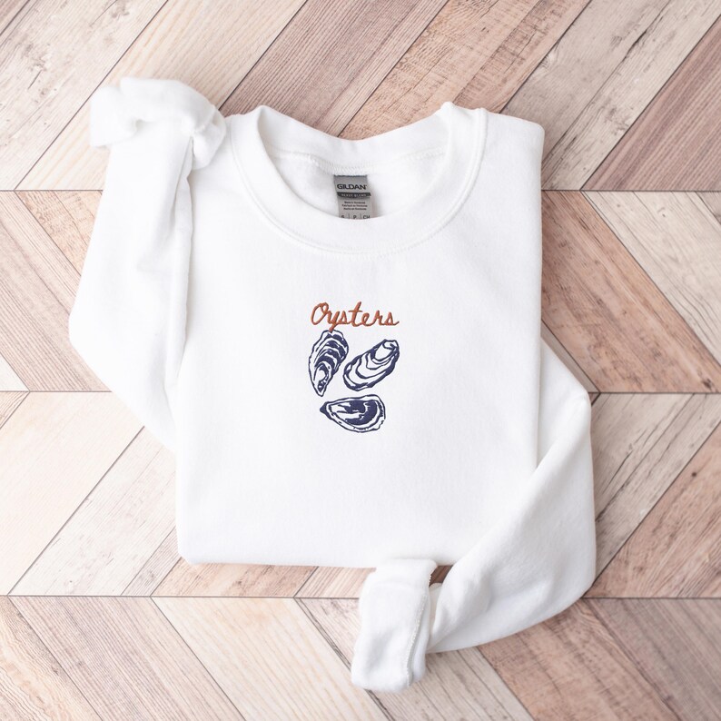 Peut inclure: Sweat-shirt blanc avec le mot "Oysters" en orange et une illustration bleue de trois hu&icirc;tres.