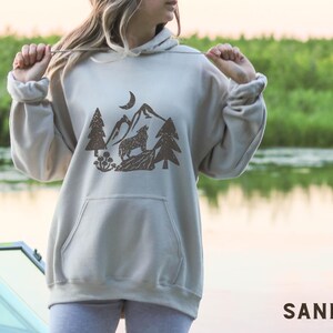 Puede incluir: Una sudadera con capucha gris claro con un estampado gr&aacute;fico marr&oacute;n de un lobo aullando a la luna en un entorno de bosque. El gr&aacute;fico incluye &aacute;rboles, monta&ntilde;as y una luna creciente.