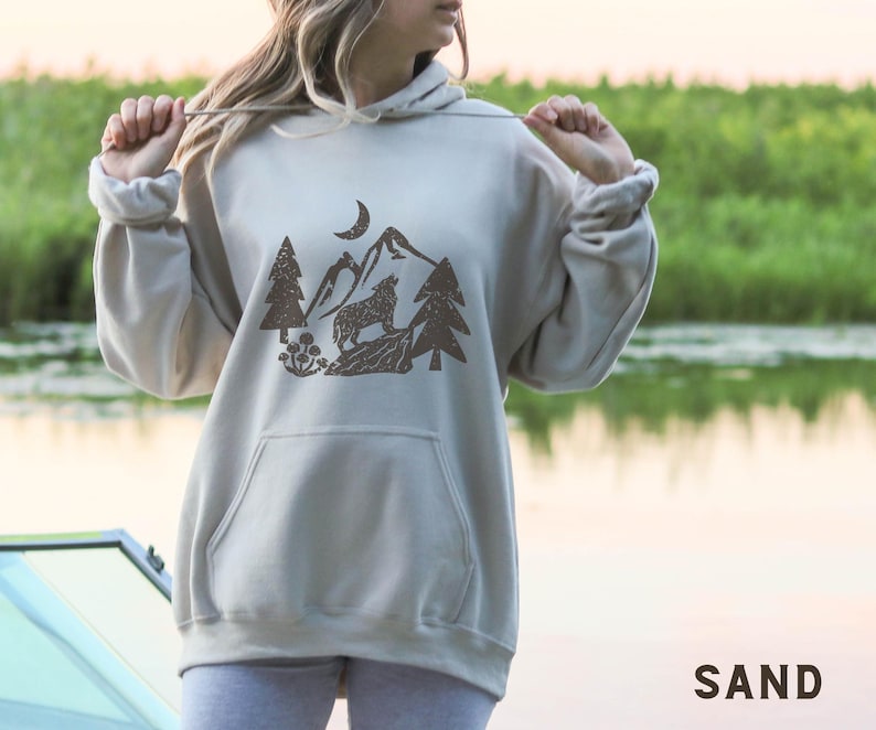 Puede incluir: Una sudadera con capucha gris claro con un estampado gr&aacute;fico marr&oacute;n de un lobo aullando a la luna en un entorno de bosque. El gr&aacute;fico incluye &aacute;rboles, monta&ntilde;as y una luna creciente.