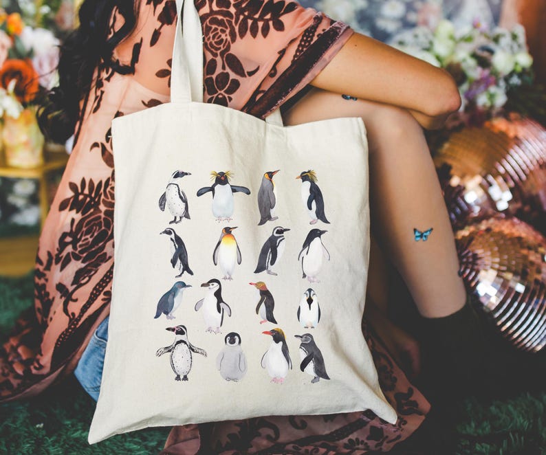 May include: Cream-colored tote bag featuring a collection of watercolor penguin illustrations. The penguins are depicted in various poses and colors, creating a whimsical and artistic design. The bag is made of a sturdy fabric.