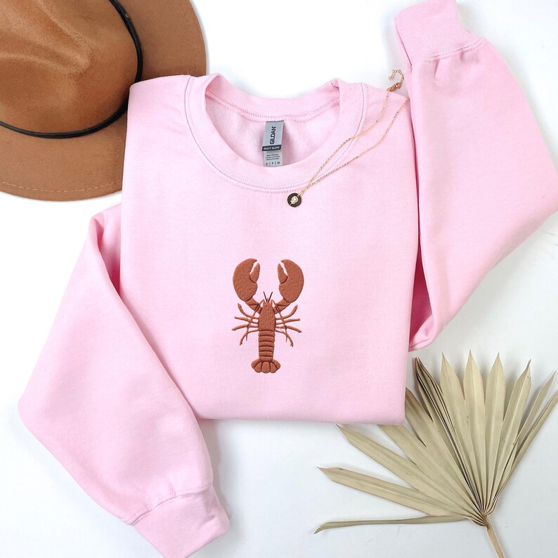 Pode incluir: Uma camisola cor-de-rosa com um lagosta castanho bordado na frente.