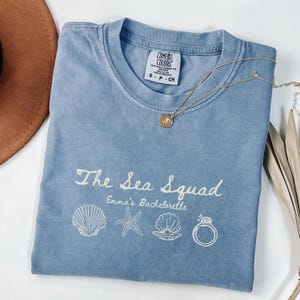 May include: A blue short-sleeve t-shirt with a crew neckline and a Comfort Colors label. The front features white text reading The Sea Squad and Emma's Bachelorette, accompanied by line art of a seashell, starfish, oyster, and diamond ring.