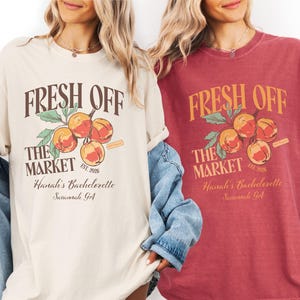 May include: Two t-shirts, one cream and one burgundy, feature the text "FRESH OFF THE MARKET" above a peach branch illustration. Below, the text reads "Hanali's Bachelorette Savannah, GA" and "EST. 2026".