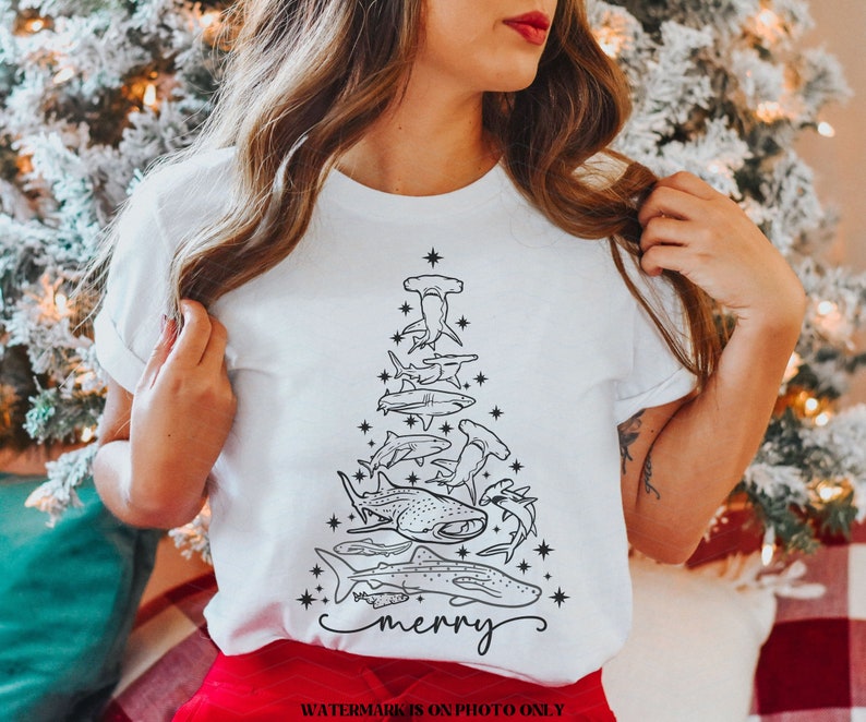 K&ouml;nnte beinhalten: Wei&szlig;es T-Shirt mit einem Weihnachtsbaum-Design aus schwarzen Hai-Illustrationen. Das Wort "merry" ist unten in Schreibschrift geschrieben. Ein einzigartiges festliches Kleidungsst&uuml;ck f&uuml;r die Weihnachtszeit.