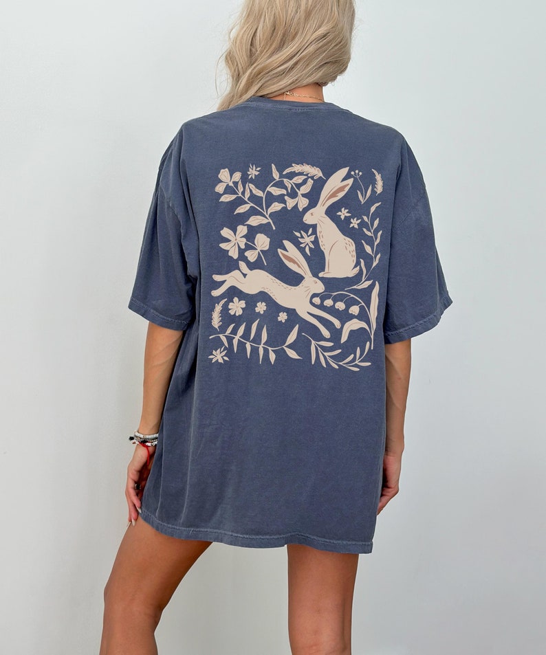 Pu&ograve; includere: Una maglietta oversize blu scuro con una grafica beige di due conigli e disegni floreali sul retro.