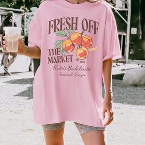 May include: A light pink t-shirt with the text "FRESH OFF THE MARKET" above a peach graphic. Below the graphic, the text reads "Katie's Bachelorette Savannah, Georgia". The shirt has short sleeves and a relaxed fit.