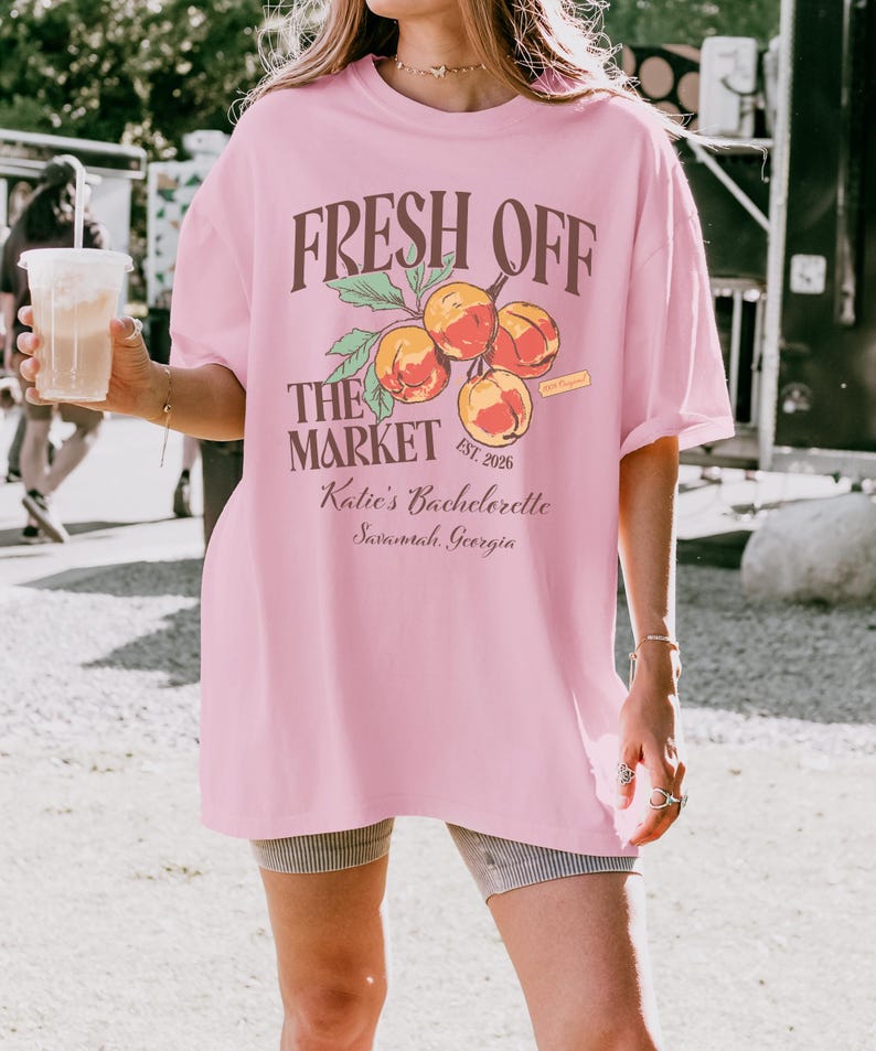 May include: A light pink t-shirt with the text "FRESH OFF THE MARKET" above a peach graphic. Below the graphic, the text reads "Katie's Bachelorette Savannah, Georgia". The shirt has short sleeves and a relaxed fit.