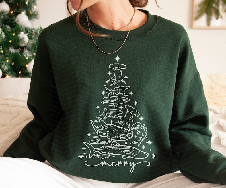 K&ouml;nnte beinhalten: Dunkelgr&uuml;nes Sweatshirt mit einem wei&szlig;en Weihnachtsbaum-Design, das aus verschiedenen Haiarten besteht. Das Wort "merry" ist unten in Schreibschrift geschrieben. Das Sweatshirt hat einen Rundhalsausschnitt und lange &Auml;rmel.