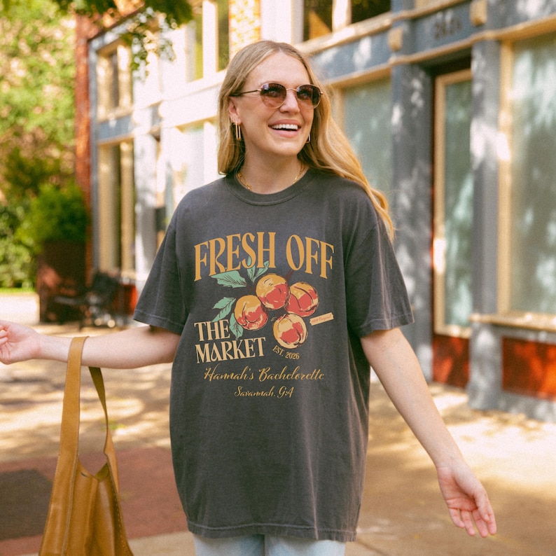 May include: Dark gray t-shirt with the text "FRESH OFF THE MARKET" and a graphic of peaches. The shirt also includes the text "Hannah's Bachelorette Savannah, GA". The shirt is a relaxed fit.