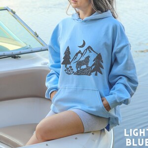Puede incluir: Una sudadera con capucha de color azul claro con un estampado gr&aacute;fico de un lobo aullando a la luna en un entorno de bosque. La sudadera tiene una capucha con cord&oacute;n y un bolsillo canguro.