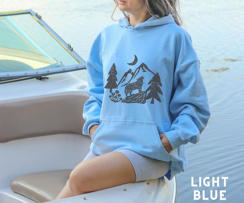 Puede incluir: Una sudadera con capucha de color azul claro con un estampado gr&aacute;fico de un lobo aullando a la luna en un entorno de bosque. La sudadera tiene una capucha con cord&oacute;n y un bolsillo canguro.