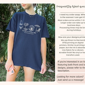 May include: A navy blue short-sleeve t-shirt featuring a white line-art graphic of a bikini, a cocktail, a pearl in an oyster shell, and a beach umbrella with a chair. The text reads Seashells and Wedding Bells and Olivia's Bachelorette.