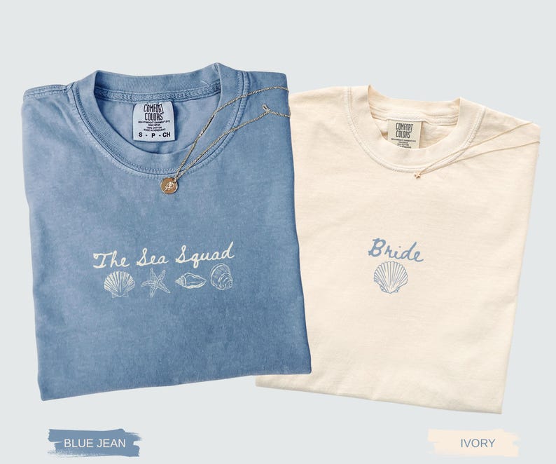 May include: A blue jean colored t-shirt with white embroidery reading The Sea Squad above four seashell icons, and an ivory t-shirt with blue embroidery reading Bride above a single seashell icon, both featuring Comfort Colors neck labels.