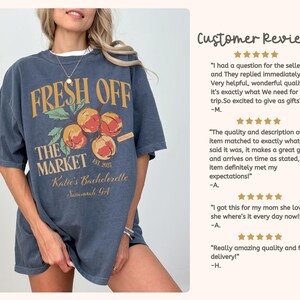 May include: A dark blue t-shirt with a graphic design featuring the text "FRESH OFF THE MARKET" above a cluster of peaches. The text "Katie's Bachelorette Savannah GA" is below the peaches.