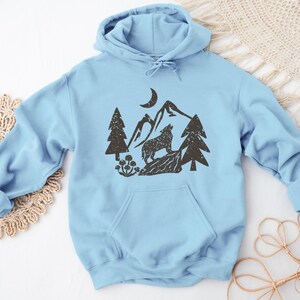 Puede incluir: Una sudadera con capucha azul claro con un estampado gr&aacute;fico negro de un lobo aullando a la luna en un paisaje de monta&ntilde;a. El gr&aacute;fico tambi&eacute;n incluye &aacute;rboles y una luna creciente.