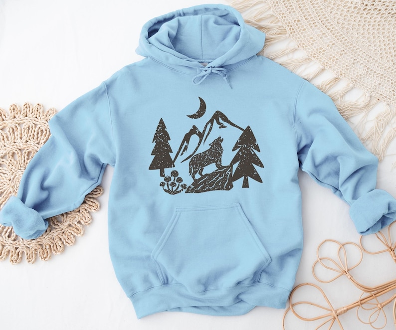 Puede incluir: Una sudadera con capucha azul claro con un estampado gr&aacute;fico negro de un lobo aullando a la luna en un paisaje de monta&ntilde;a. El gr&aacute;fico tambi&eacute;n incluye &aacute;rboles y una luna creciente.