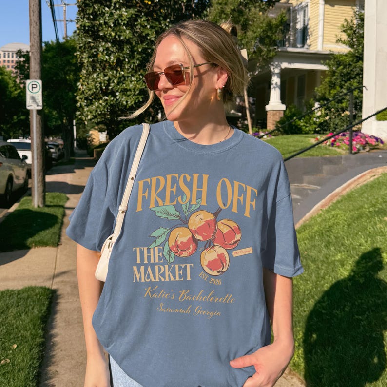 May include: A blue t-shirt with the text "FRESH OFF THE MARKET" and a graphic of peaches. The shirt also includes the text "Katie's Bachelorette Savannah, Georgia".