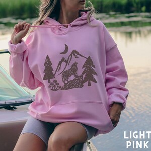 Puede incluir: Una sudadera con capucha rosa claro con un gr&aacute;fico marr&oacute;n de un lobo aullando a la luna en un paisaje de monta&ntilde;a. El gr&aacute;fico tambi&eacute;n incluye &aacute;rboles y flores.
