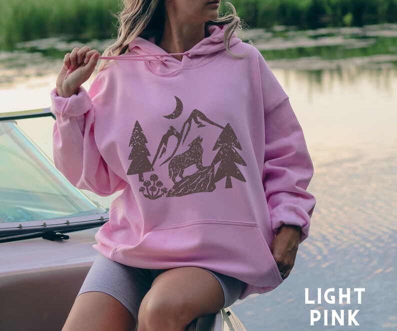 Puede incluir: Una sudadera con capucha rosa claro con un gr&aacute;fico marr&oacute;n de un lobo aullando a la luna en un paisaje de monta&ntilde;a. El gr&aacute;fico tambi&eacute;n incluye &aacute;rboles y flores.