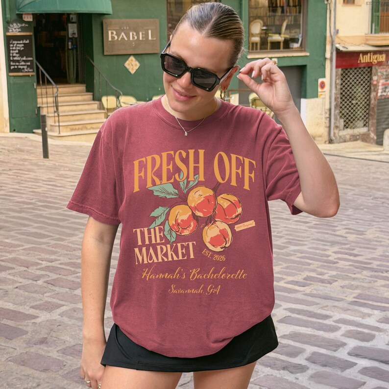 May include: A maroon t-shirt with the text "FRESH OFF THE MARKET" in gold lettering, along with a graphic of peaches. The shirt also includes the text "Hannah's Bachelorette Savannah, GA" and "EST. 2026".
